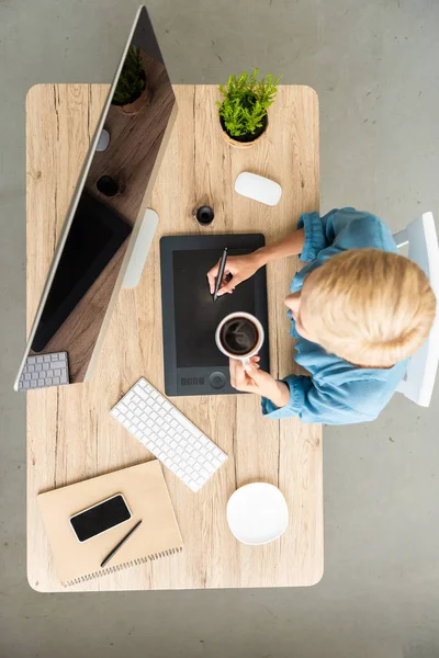 Над головой вид женщины-фрилансера, пьющей кофе, работающей на графическом планшете за столом со смартфоном и компьютером в домашнем офисе — стоковое фото