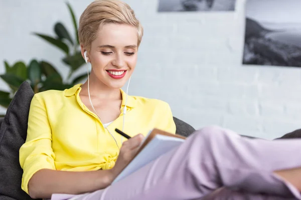 Smiling young woman in earphones writing in textbook on sofa at home — Stock Photo