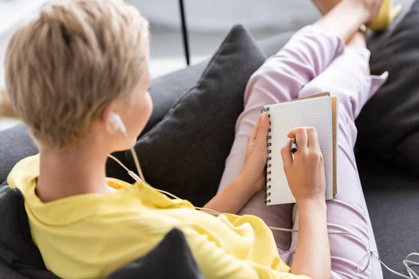 Rückansicht einer jungen Frau mit Kopfhörern, die in einem Lehrbuch auf dem heimischen Sofa schreibt — Stockfoto