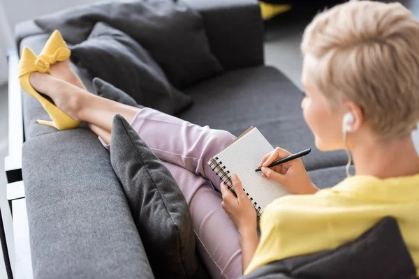 Selective focus of stylish woman in earphones writing in textbook on sofa at home — Stock Photo