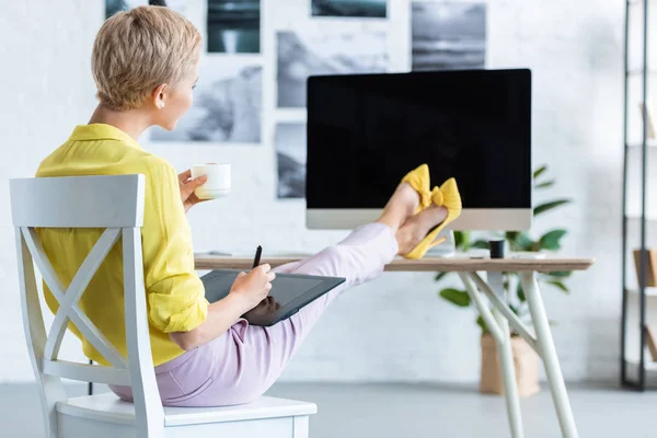 Selective focus of female freelancer drinking coffee and using graphic tablet at table with computer at home office — Stock Photo