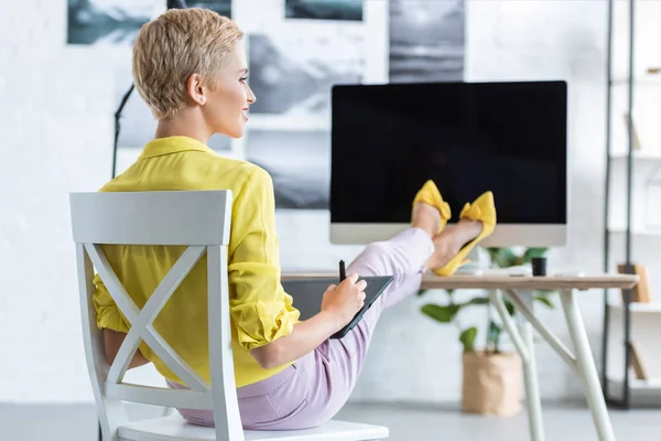 Sonriente freelancer femenina trabajando en tableta gráfica en la mesa con la computadora en la oficina en casa - foto de stock