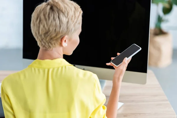 Junge Geschäftsfrau checkt Smartphone am Tisch mit Computer im Büro — Stockfoto