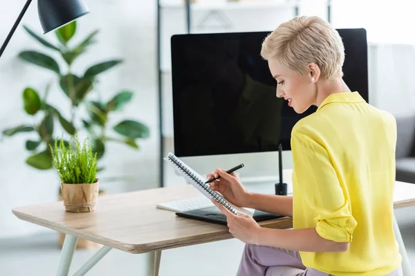 Sorrindo freelancer feminino escrevendo em livro didático à mesa com tablet gráfico e computador — Fotografia de Stock