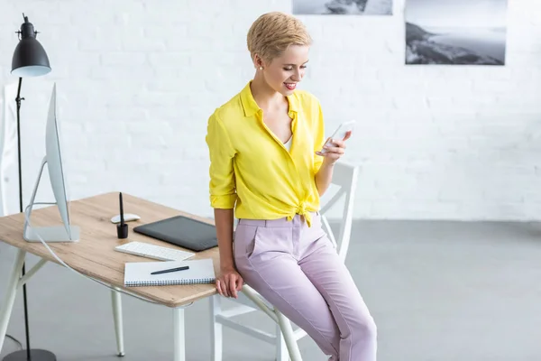 Elegante freelancer femenina sonriente usando smartphone cerca de la mesa con tableta gráfica y computadora en la oficina en casa - foto de stock