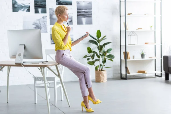 Vista lateral del pensativo freelancer femenino con bolígrafo bajo la barbilla que sostiene el libro de texto en la oficina en casa - foto de stock