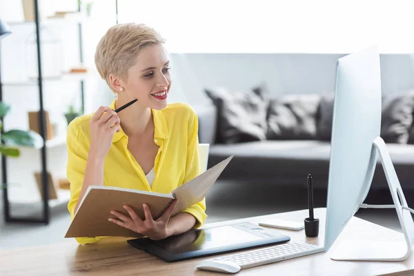 Pensativo freelancer femenino sosteniendo libro de texto y sentado a la mesa con computadora y tableta gráfica - foto de stock