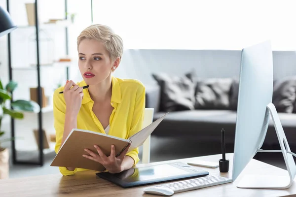 Pensive female freelancer holding textbook and sitting at table with computer and graphic tablet — Stock Photo