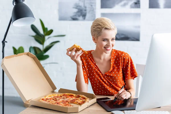 Happy female freelancer eating pizza and drawing on graphic tablet at home office — Stock Photo