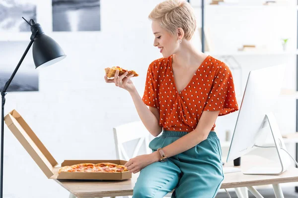 Side view of smiling female freelancer sitting on table and eating pizza at home office — Stock Photo