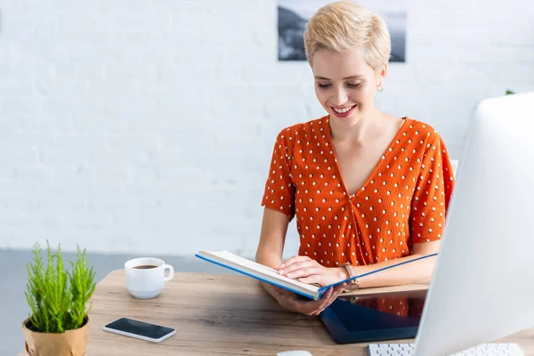 Libro di lettura femminile freelance sorridente a tavola con tablet grafico e computer in home office — Foto stock