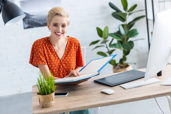 Female freelancer holding book at table with graphic tablet and computer in home office — Stock Photo