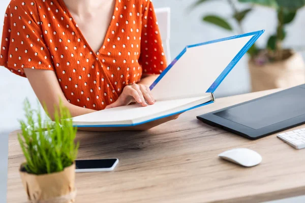 Cropped image of female freelancer reading book at table with graphic tablet, smartphone and computer in home office — Stock Photo