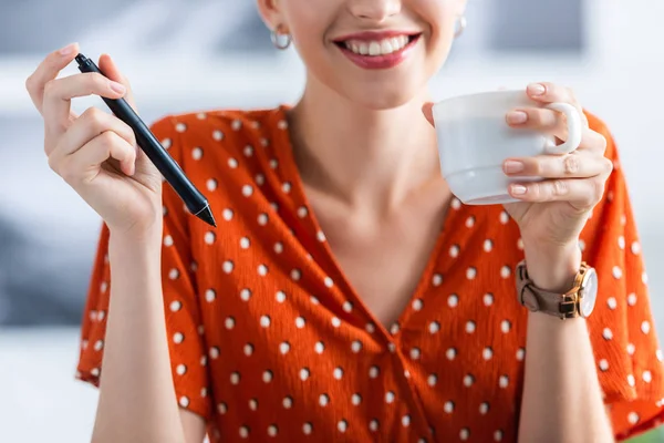 Частковий вид усміхненого фрілансера жінки з чашкою кави, що тримає ручку для графічного планшета в домашньому офісі — стокове фото