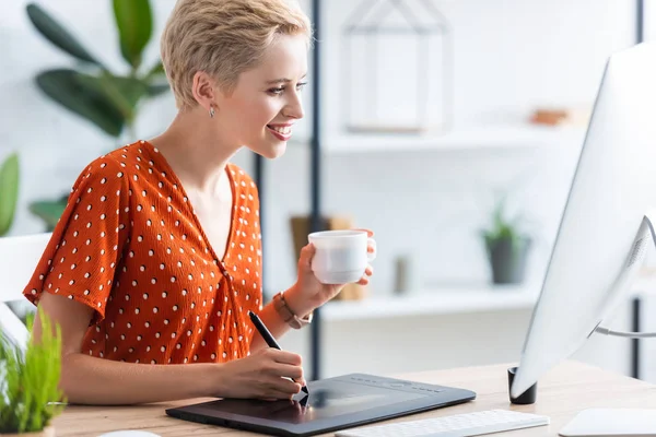 Vista lateral de la mujer freelancer feliz con el dibujo de taza de café en la tableta gráfica en la mesa con el ordenador - foto de stock