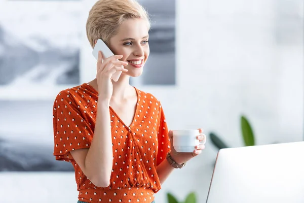 Усміхнена бізнес-леді розмовляє на смартфоні і тримає чашку кави біля настільного комп'ютера в офісі — Stock Photo