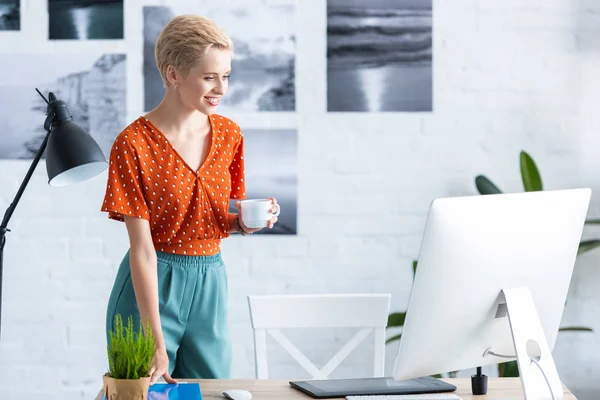 Freelancer feminino sorrindo segurando xícara de café perto da mesa com tablet gráfico e computador no escritório em casa — Fotografia de Stock