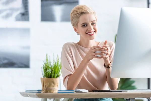 Smiling businesswoman holding cup of coffee at table with graphic tablet and computer in office — Stock Photo