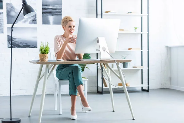Feliz empresaria sosteniendo la taza de café en la mesa con la tableta gráfica y el ordenador en la oficina - foto de stock