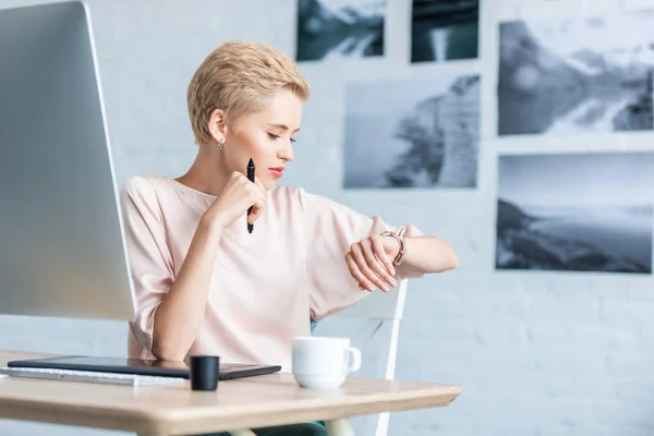 Freelancer feminino olhando para relógio de pulso à mesa com tablet gráfico e computador em casa escritório — Fotografia de Stock