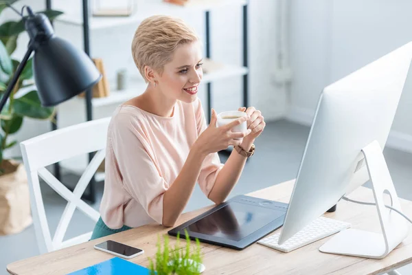 Улыбающаяся женщина-фрилансер пьет кофе за столом с графическим планшетом и компьютером в домашнем офисе — стоковое фото