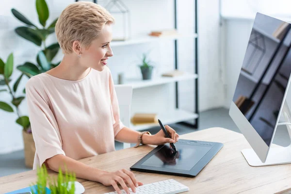 Seitenansicht einer lächelnden Freiberuflerin, die am Tisch im Home Office auf einem Grafik-Tablet zeichnet — Stockfoto