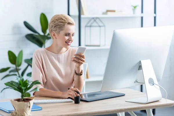 Smiling female freelancer holding smartphone at table with graphic tablet and computer in home office — Stock Photo