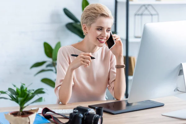 Sorrindo fotógrafo feminino falando no smartphone e usando tablet gráfico no escritório em casa — Fotografia de Stock