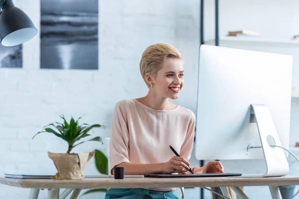 Smiling female freelancer drawing on graphic tablet at table with computer — Stock Photo