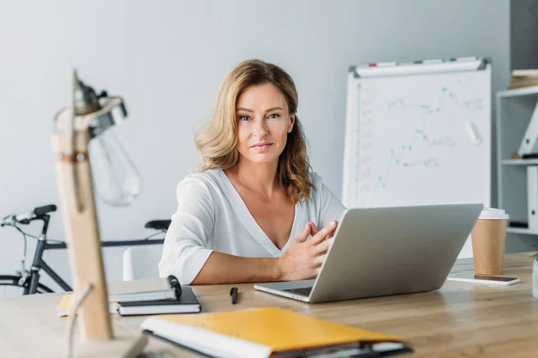 Attraktive Geschäftsfrau sitzt mit Laptop am Tisch im Büro und blickt in die Kamera — Stockfoto