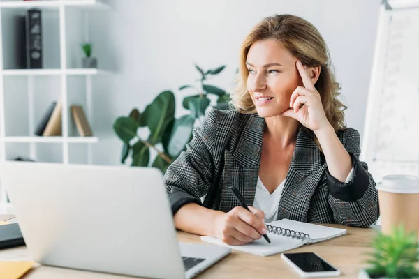 Задумчивая привлекательная деловая женщина сидит за столом с ноутбуком и ноутбуком в офисе — стоковое фото