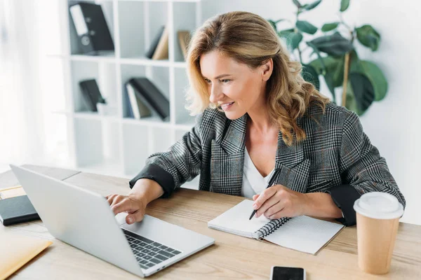 Attractive businesswoman using laptop and writing something to notebook in office — Stock Photo