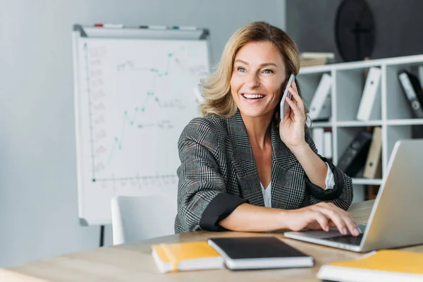 Sonriente hermosa mujer de negocios hablando por teléfono inteligente en la oficina y mirando hacia otro lado - foto de stock