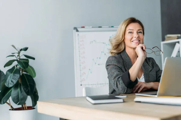 Chère femme d'affaires souriante tenant des lunettes et regardant loin dans le bureau — Photo de stock