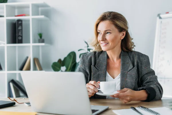Beautiful businesswoman sitting with cup of coffee at table in office and looking away — Stock Photo