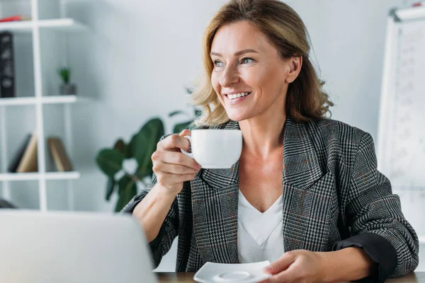 Приваблива бізнес-леді, сидячи з чашкою кави за столом в офісі і дивлячись далеко — стокове фото
