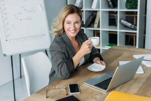 Attractive businesswoman sitting with cup of coffee at table in office — Stock Photo