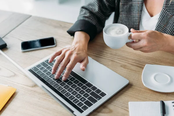 Обрезанный образ бизнесвумен с помощью ноутбука и проведение чашки кофе в офисе — стоковое фото
