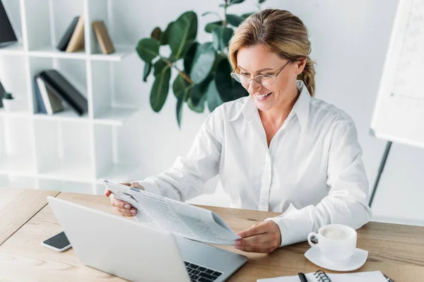 Усміхнена приваблива бізнес-леді читає газету за столом з ноутбуком в офісі — стокове фото
