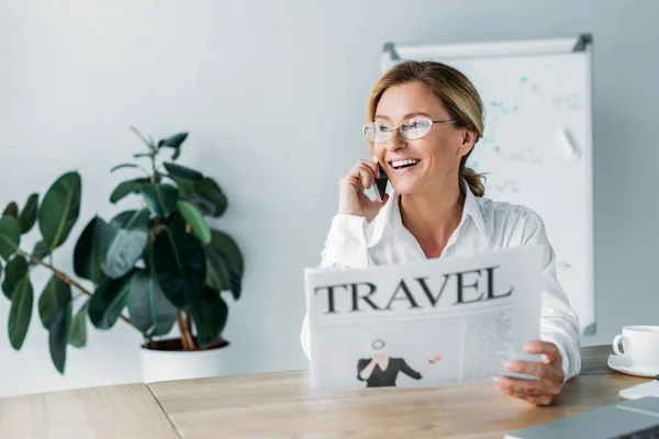 Sonriente atractiva mujer de negocios hablando por teléfono inteligente y sosteniendo periódico de viaje en la oficina - foto de stock