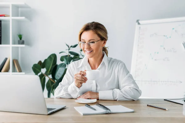 Attractive businesswoman holding cup of coffee and looking at laptop in office — Stock Photo