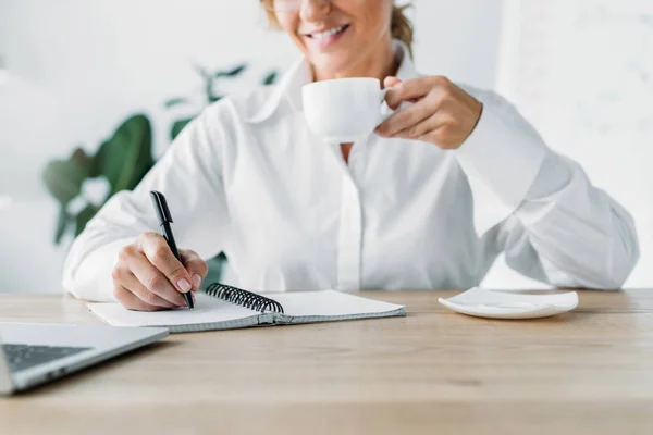 Image recadrée femme d'affaires tenant tasse de café et l'écriture au carnet dans le bureau — Photo de stock