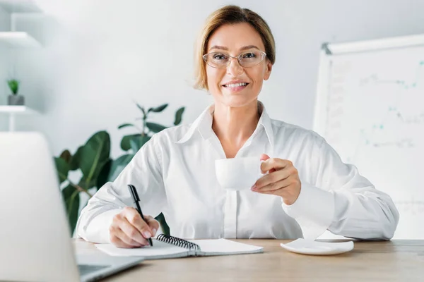 Attraktive Geschäftsfrau hält Tasse Kaffee in der Hand und schreibt im Büro ans Notizbuch — Stockfoto