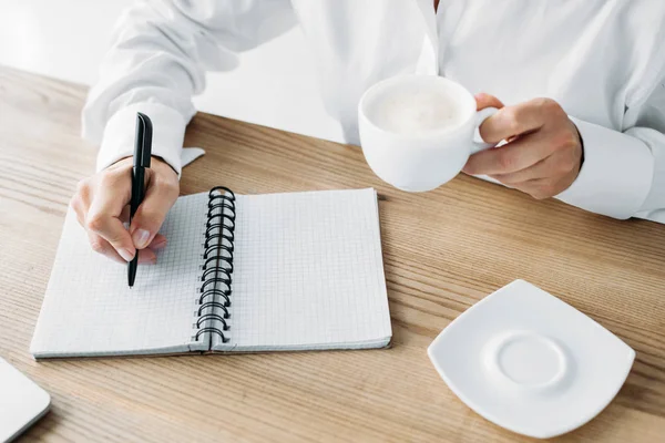 Обрізана бізнес-леді тримає чашку кави і пише в блокноті в офісі — стокове фото