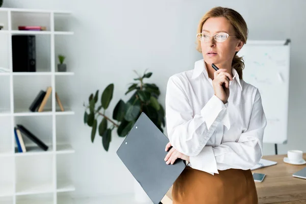 Attractive businesswoman holding clipboard and looking away in office — Stock Photo