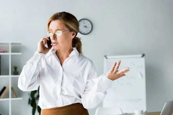 Attractive businesswoman talking by smartphone and gesturing in office — Stock Photo