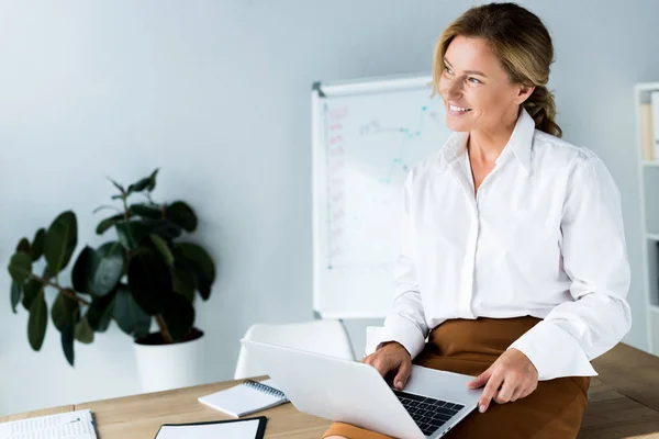 Attractive businesswoman sitting on table with laptop and looking away in office — Stock Photo
