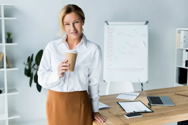 Beautiful businesswoman holding disposable coffee cup in office and looking at camera — Stock Photo