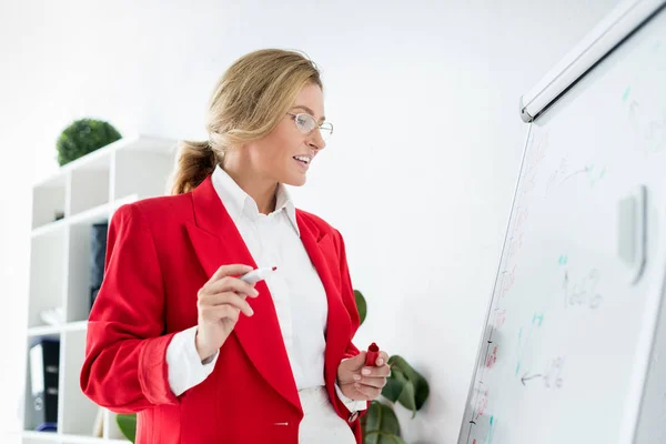 Attractive businesswoman in red jacket standing near flipchart in office and holding marker — Stock Photo