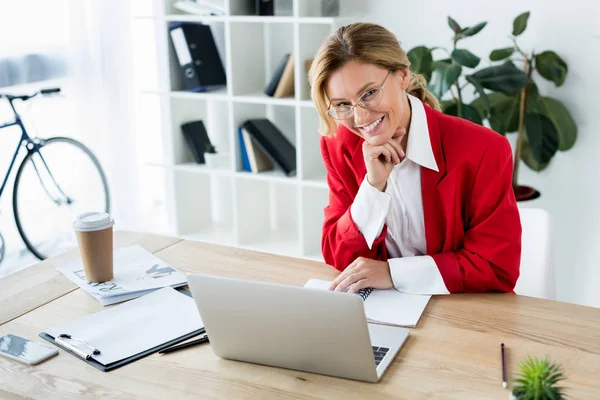 Atractiva mujer de negocios sonriente sentado en la mesa con el ordenador portátil en la oficina y mirando a la cámara - foto de stock